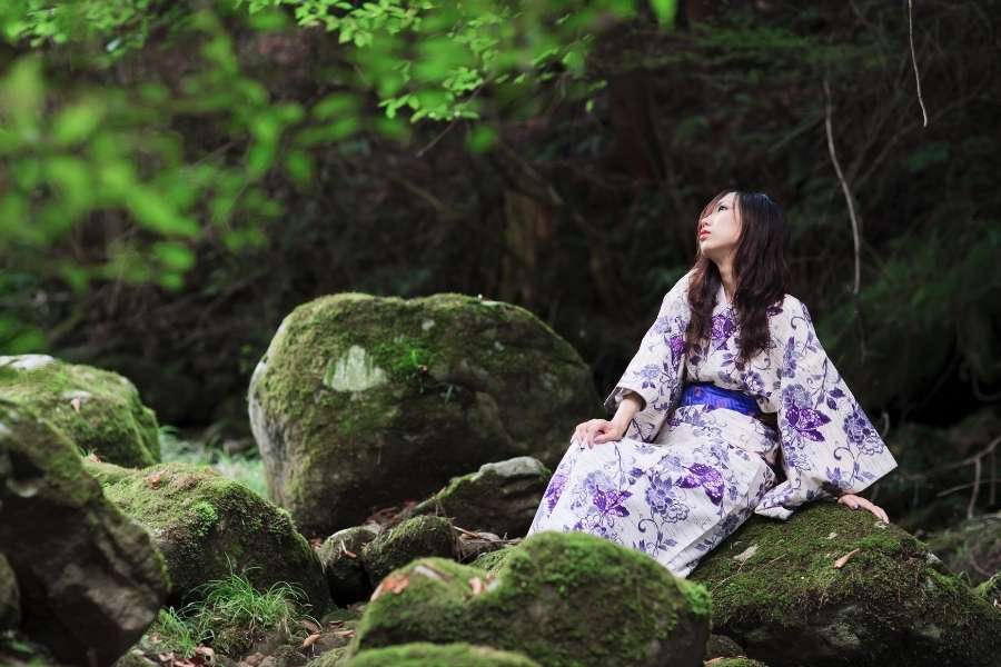 Une jeune femme en kimono violet et blanc est assise en contemplation sur un rocher couvert de mousse dans une forêt profonde.