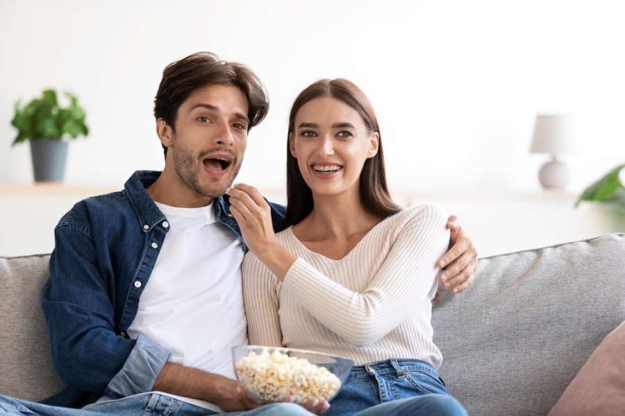 Un couple surpris sur un canapé, l'homme la bouche ouverte et la femme souriante, partageant un bol de pop-corn.